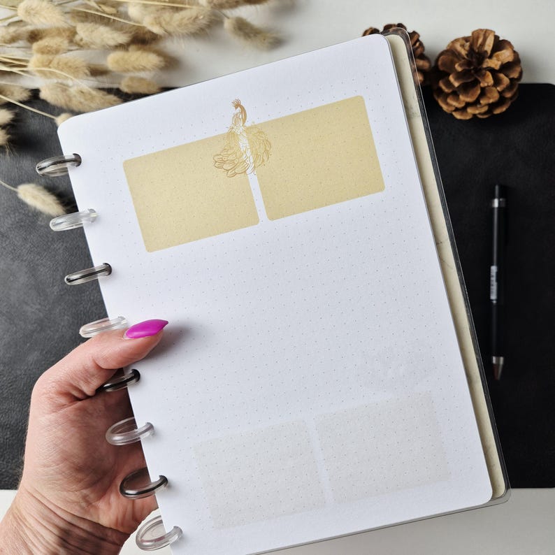 May include: A disc-bound notebook with white dot-grid paper featuring two rectangular light yellow adhesive notes at the top, centered with a gold-colored line drawing of a person in a flowing dress, and two faint translucent rectangular stickers below.