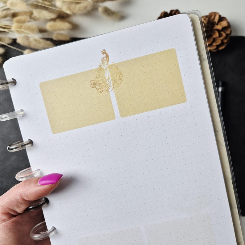 May include: A white dot grid planner page featuring two rectangular light yellow adhesive labels placed side by side. A gold line art illustration of a peacock is centered above the gap between the two rectangular labels.