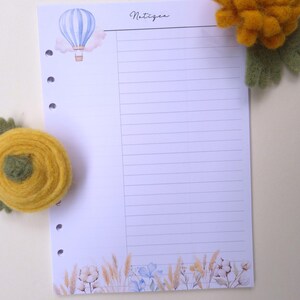 Op de afbeelding: Gelijnd papier met een aquarel illustratie van een luchtballon en bloemmotieven. Het woord "Notizen" is in cursief geschreven. Twee gele vilten bloemdecoraties zijn boven en onder het papier geplaatst.