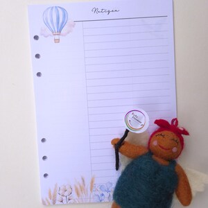 May include: A lined planner page with a watercolor hot air balloon illustration and the word "Notizen" in script. A felt doll with a sign is in the foreground. The page has three holes for binding.