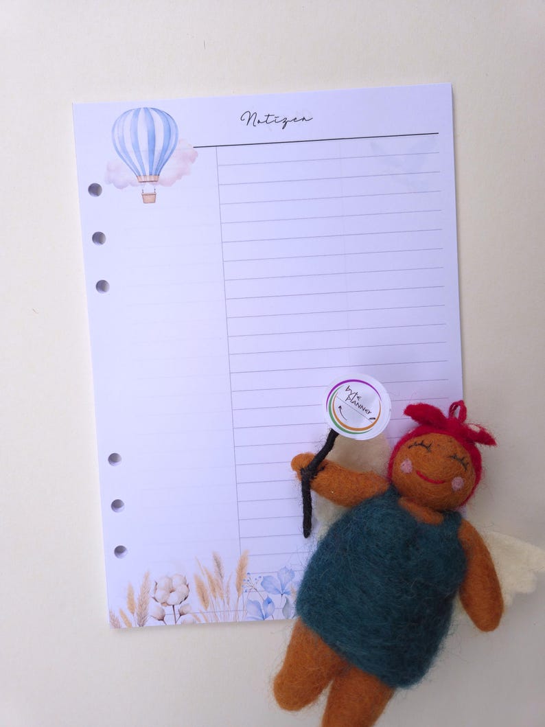 May include: A lined planner page with a watercolor hot air balloon illustration and the word "Notizen" in script. A felt doll with a sign is in the foreground. The page has three holes for binding.