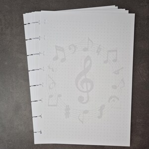 May include: A stack of white dot grid paper with a musical theme. The pages feature a light grey design of musical notes and symbols, including a treble clef. The paper is bound on the left side with black binding.