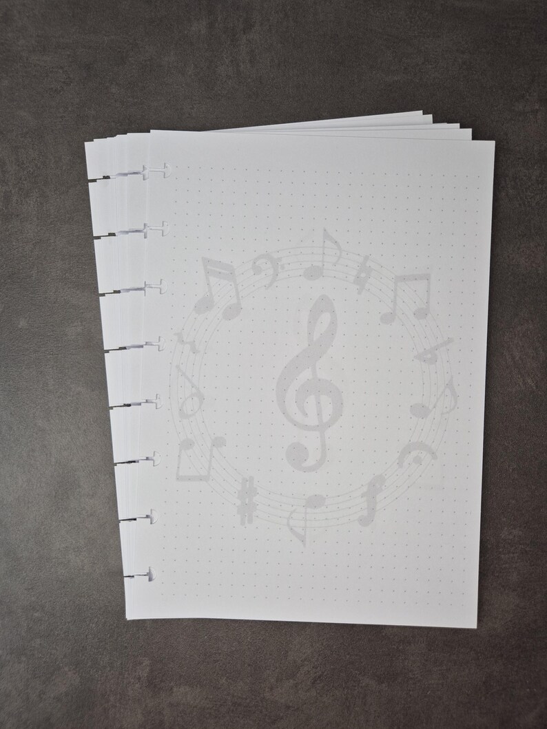 May include: A stack of white dot grid paper with a musical theme. The pages feature a light grey design of musical notes and symbols, including a treble clef. The paper is bound on the left side with black binding.