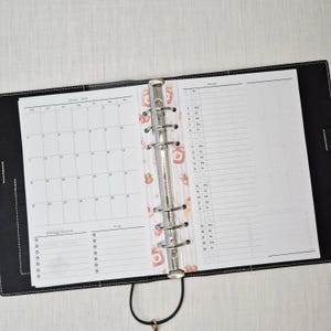 May include: Open black leather binder with a silver metal ring mechanism. The binder is open to reveal a monthly calendar and a daily planner page. A decorative strawberry-themed divider is visible.