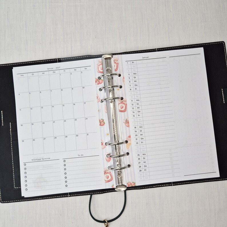May include: Open black leather binder with a silver metal ring mechanism. The binder is open to reveal a monthly calendar and a daily planner page. A decorative strawberry-themed divider is visible.