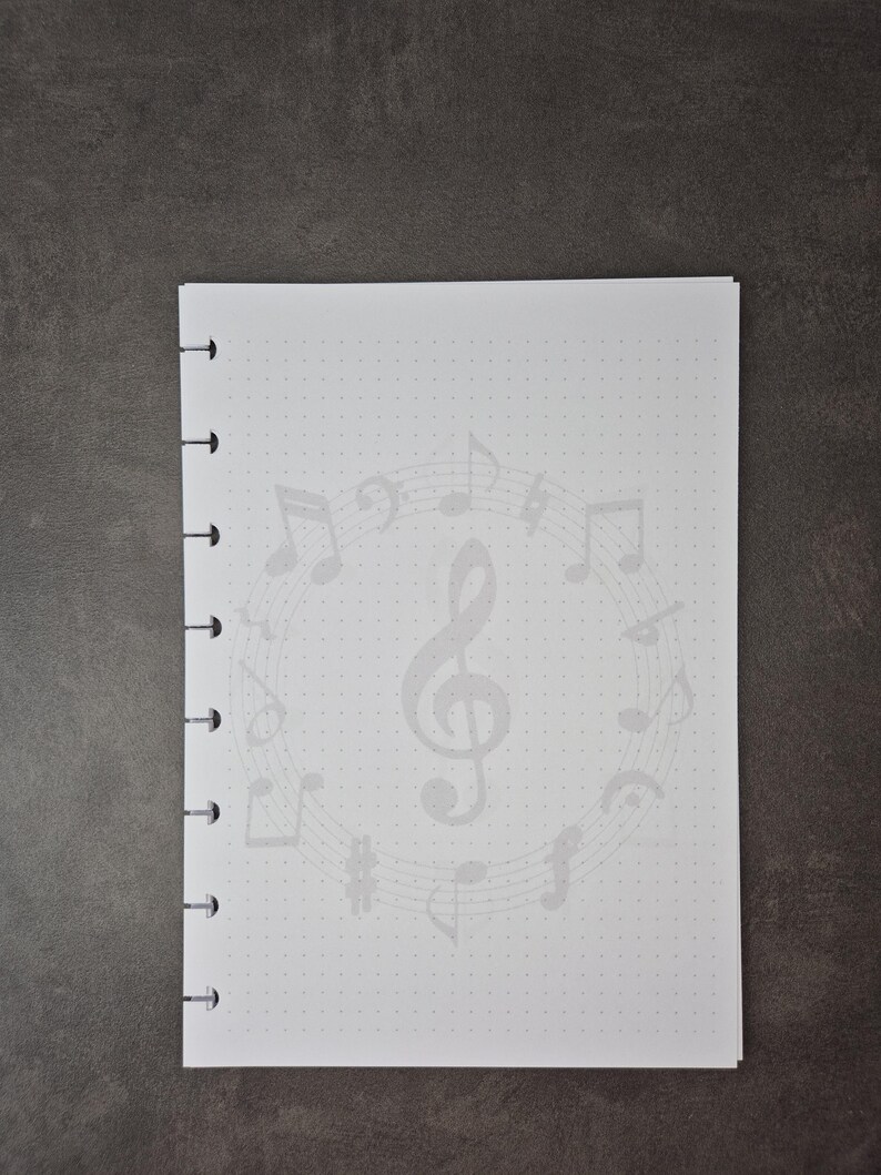May include: A white notebook with a musical theme. The notebook features a light grey design of musical notes and a treble clef. The pages have a dotted grid pattern. The notebook is bound with black rings.