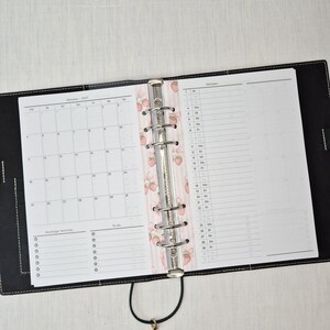 May include: Black leather binder open to a calendar and to-do list. The calendar is for October 2021. A decorative pink and white strawberry pattern is visible on the binder's spine. The binder is open on a white surface.