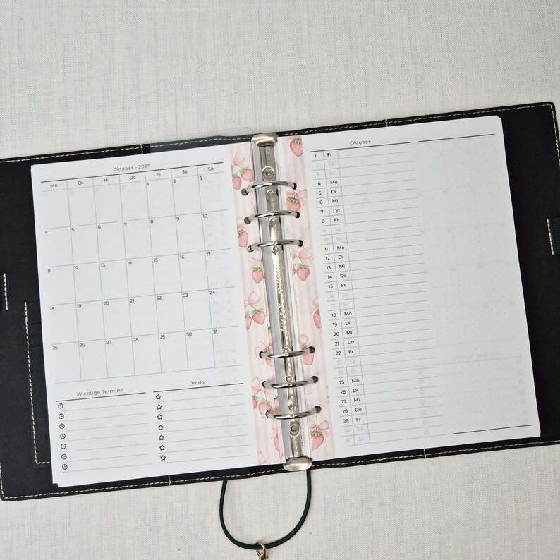 May include: Black leather binder open to a calendar and to-do list. The calendar is for October 2021. A decorative pink and white strawberry pattern is visible on the binder's spine. The binder is open on a white surface.