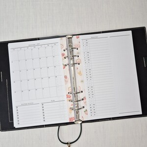 May include: Open black leather binder with a calendar and lined pages. The calendar shows the month of December 2021. A decorative strip with strawberries and ice cream separates the calendar from the lined pages. A black cord with a gold-colored charm is attached.