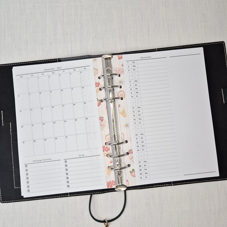 May include: Open black leather binder with a calendar and lined pages. The calendar shows the month of December 2021. A decorative strip with strawberries and ice cream separates the calendar from the lined pages. A black cord with a gold-colored charm is attached.