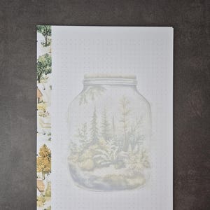 May include: A notebook with a watercolor illustration of a forest scene inside a jar. The notebook has a dotted grid pattern on the pages. The left side of the notebook has a decorative border with trees and houses.