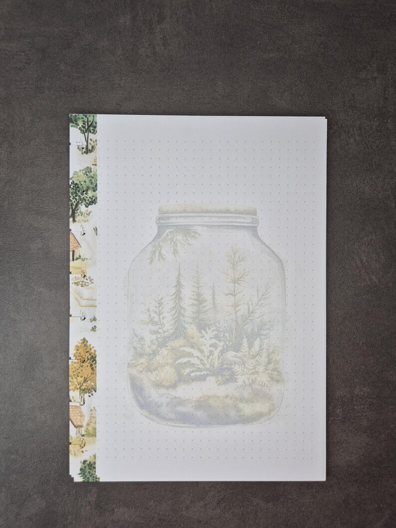 May include: A notebook with a watercolor illustration of a forest scene inside a jar. The notebook has a dotted grid pattern on the pages. The left side of the notebook has a decorative border with trees and houses.