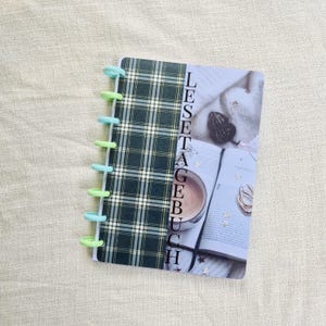 Peut inclure: Un petit carnet avec une couverture à carreaux verts et noirs et le mot "LESETAGBUCH" imprimé verticalement. Le carnet a une reliure spirale avec des anneaux bleu clair et verts. Le côté droit de la couverture présente une photo d'un livre, d'un café et d'objets décoratifs.