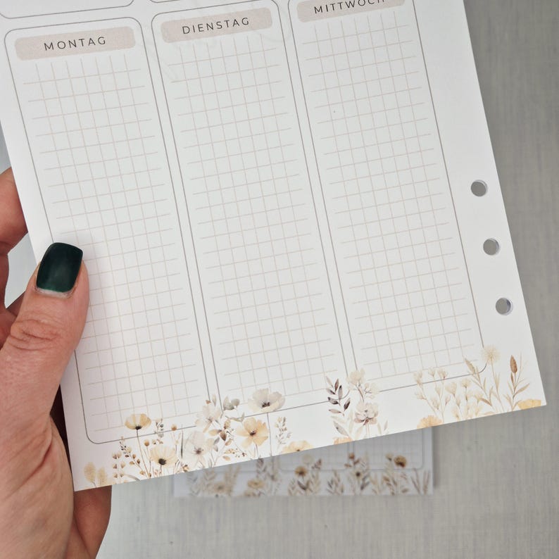May include: A weekly planner page with a grid layout for scheduling. The top of the page has the days of the week in German. The bottom of the page features a floral design in neutral tones. The page is held by a hand with a dark green nail polish.