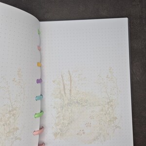 May include: Open notebook with dot grid pages and a watercolor illustration of a path with paw prints. The notebook has colorful plastic disc binding and a soft, neutral color palette.