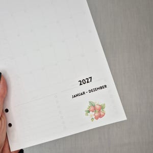 May include: A white 2027 calendar page with the text "JANUAR - DEZEMBER" and a strawberry illustration. The calendar has a grid layout for monthly planning and is held by a hand.