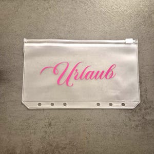 Peut inclure: Pochette transparente avec fermeture éclair. Le mot "Urlaub" est écrit en rose sur le devant. La pochette comporte six trous le long du bord inférieur.