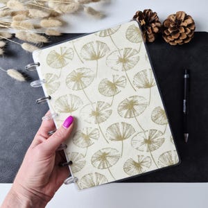 May include: A disc-bound notebook featuring a cream-colored cover with a repeating pattern of gold-toned, fan-shaped botanical illustrations. The notebook has clear plastic discs along the spine and a protective transparent cover layer.