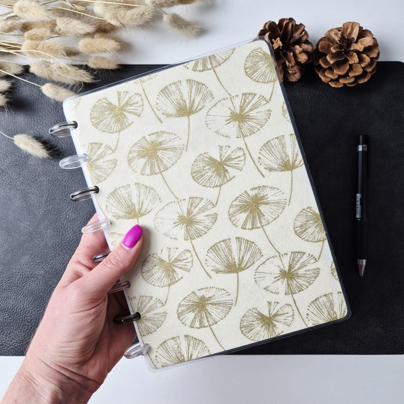 May include: A disc-bound notebook featuring a cream-colored cover with a repeating pattern of gold-toned, fan-shaped botanical illustrations. The notebook has clear plastic discs along the spine and a protective transparent cover layer.