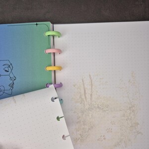 May include: Open notebook with colorful plastic rings. The left page has a gradient of blue and green with a line drawing of a face. The right page has a dotted grid and a faded illustration of a forest scene with paw prints.