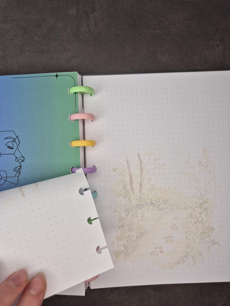 May include: Open notebook with colorful plastic rings. The left page has a gradient of blue and green with a line drawing of a face. The right page has a dotted grid and a faded illustration of a forest scene with paw prints.