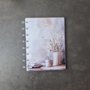 May include: A small, clear-bound notebook with a watercolor-style cover. The cover art depicts a still life with a vase of dried flowers, a smaller container with pencils, and an open book. The color palette is soft, with muted tones of beige, gray, and white.
