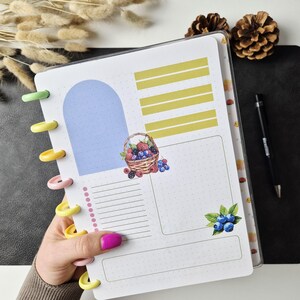 May include: A disc-bound planner page featuring a light blue arch, yellow horizontal bars, and a watercolour illustration of a basket filled with berries. A small cluster of blueberries with green leaves is at the bottom right, alongside a dotted list.
