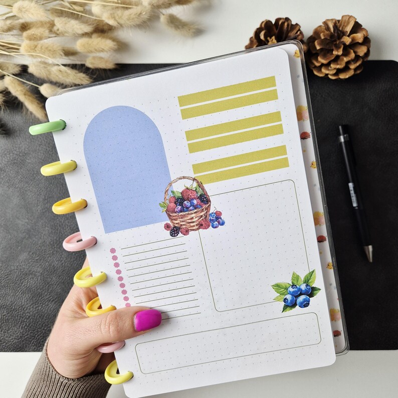 May include: A disc-bound planner page featuring a light blue arch, yellow horizontal bars, and a watercolour illustration of a basket filled with berries. A small cluster of blueberries with green leaves is at the bottom right, alongside a dotted list.
