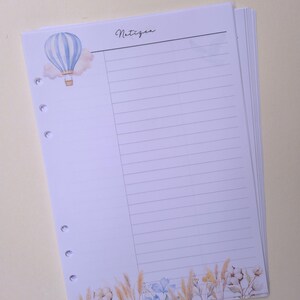 May include: A stack of lined paper with a watercolor design. The top sheet features a blue and white hot air balloon, the word "Notizen", and a floral border with beige and blue accents. The paper is hole-punched for a binder.