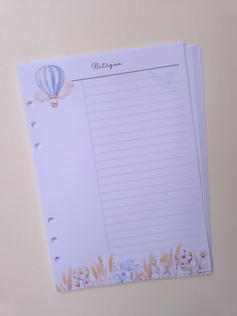 May include: A stack of lined paper with a watercolor design. The top sheet features a blue and white hot air balloon, the word "Notizen", and a floral border with beige and blue accents. The paper is hole-punched for a binder.