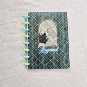 Peut inclure: Un petit journal à spirales avec une couverture à motifs géométriques vert d'eau et dorés. La couverture présente un motif de fenêtre cintrée avec un arrangement floral et des livres ouverts. Le mot "Lesejournal" est écrit en cursive.