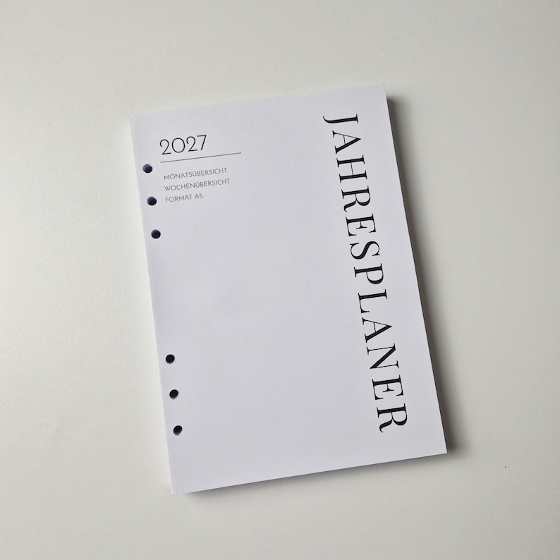 K&ouml;nnte beinhalten: Ein wei&szlig;er A5-Jahresplaner f&uuml;r das Jahr 2027 mit schwarzem Text. Das Deckblatt zeigt vertikal das Wort JAHRESPLANER sowie 2027, MONATS&Uuml;BERSICHT, WOCHEN&Uuml;BERSICHT und FORMAT A5. Die linke Seite ist mit sechs runden Lochungen f&uuml;r eine Bindung versehen.