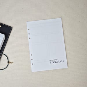 May include: A white planner refill page with six punched holes along the left edge. The page features a layout of empty rectangular boxes and the printed text w&ouml;chentlicher R&Uuml;CKBLICK. A black binder corner is visible on the left side.