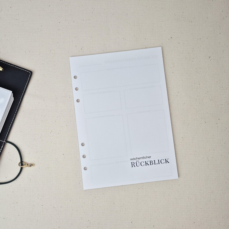 May include: A white planner refill page with six punched holes along the left edge. The page features a layout of empty rectangular boxes and the printed text w&ouml;chentlicher R&Uuml;CKBLICK. A black binder corner is visible on the left side.