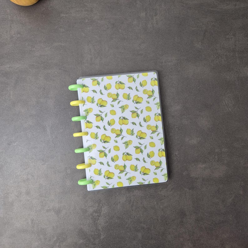 Könnte beinhalten: Ein kleines Notizbuch mit durchsichtiger Kunststoffhülle und Spiralbindung. Das Cover zeigt ein Muster aus gelben Zitronen und grünen Blättern auf weißem Hintergrund. Die Spiralbindung hat abwechselnd grüne und gelbe Ringe.
