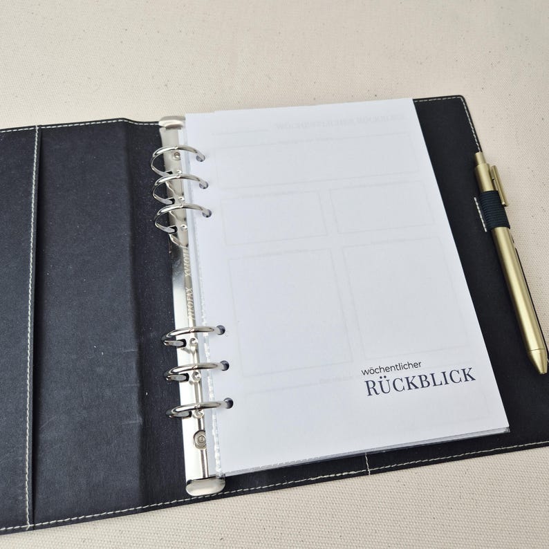 May include: A black six-ring binder with white stitching, containing white paper inserts with the printed text w&ouml;chentlicher R&Uuml;CKBLICK. A gold-toned metal pen with a black grip is secured in a loop on the right side of the open binder cover.