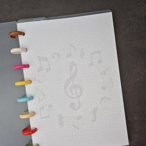 May include: Open notebook with a white dotted page featuring a musical note design. The notebook has colourful plastic rings in brown, white, pink, yellow, blue, beige, red, and green. The notebook is open to a blank page.
