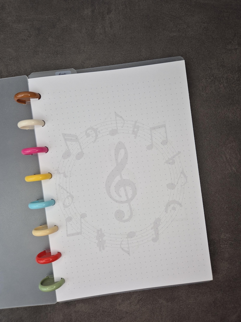 May include: Open notebook with a white dotted page featuring a musical note design. The notebook has colourful plastic rings in brown, white, pink, yellow, blue, beige, red, and green. The notebook is open to a blank page.