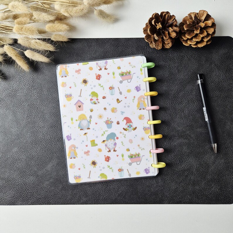 May include: A disc-bound notebook featuring a white cover with a repeating pattern of colourful garden gnomes, flowers, watering cans, and wheelbarrows. The notebook has pastel-coloured plastic discs along the side and is accompanied by a black pen.