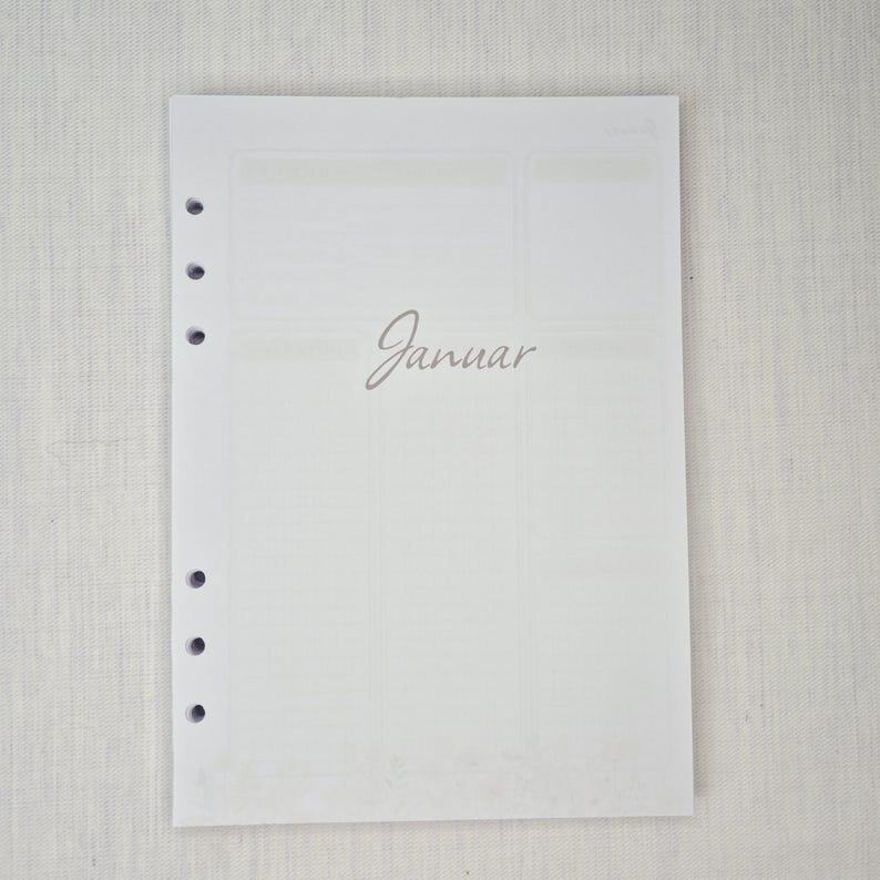 May include: A white planner page with the word "Januar" in a cursive font. The page has a grid layout with sections for notes and scheduling. The page has several holes on the left side for binding.