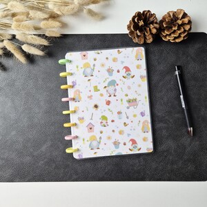 May include: A disc-bound notebook featuring a white cover with a repeating pattern of garden gnomes, birdhouses, flowers, and gardening tools. The notebook has colourful plastic discs along the left side and is accompanied by a black ballpoint pen.