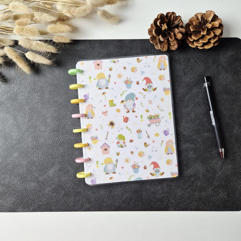 May include: A disc-bound notebook featuring a white cover with a repeating pattern of garden gnomes, birdhouses, flowers, and gardening tools. The notebook has colourful plastic discs along the left side and is accompanied by a black ballpoint pen.