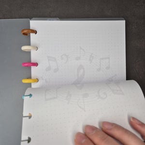May include: A grey notebook with coloured plastic rings. The open pages display a dotted grid and a faint musical note design. A hand turns a page, showing the notebook's interior.