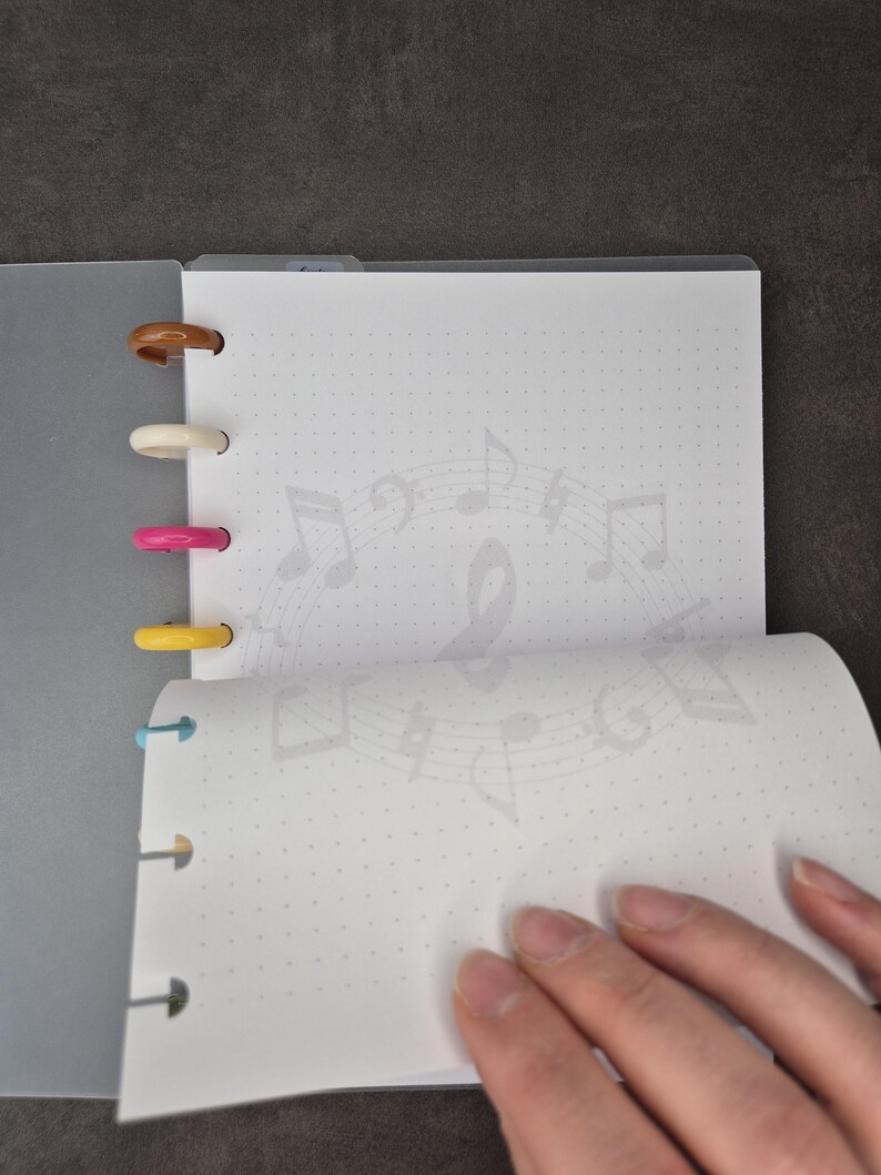 May include: A grey notebook with coloured plastic rings. The open pages display a dotted grid and a faint musical note design. A hand turns a page, showing the notebook's interior.