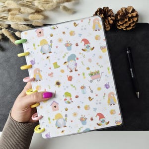 May include: A disc-bound notebook with a white cover featuring a repeating pattern of garden gnomes, flowers, watering cans, and birdhouses. The notebook has colourful plastic binding discs in yellow, green, and pink along the left side edge.
