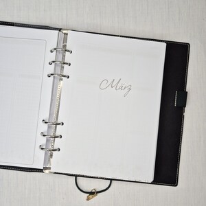 May include: Open black leather binder with silver metal rings and a clasp. The word "M&auml;rz" is written in cursive on a white page. The binder is open, revealing blank pages and a clear plastic divider.