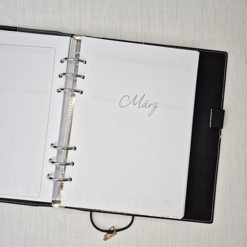 May include: Open black leather binder with silver metal rings and a clasp. The word "M&auml;rz" is written in cursive on a white page. The binder is open, revealing blank pages and a clear plastic divider.