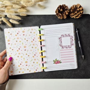 May include: A disc-bound planner featuring a left page with a pattern of small yellow flowers, green leaves, and tiny bees, and a right page with lined sections, a dotted box, a raspberry wreath graphic, and a raspberry illustration.