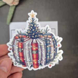Könnte beinhalten: Ein Aufkleber mit einem Stapel offener Bücher, die als Weihnachtsschmuck dekoriert sind. Ein Weihnachtsbaum sitzt auf den Büchern, geschmückt mit einem Stern, Lichtern und Kugeln. Die Bücher haben rote, blaue und goldene Buchrücken.