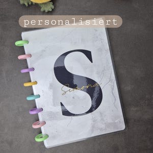 Puede incluir: Un cuaderno personalizado con una cubierta con efecto mármol, con una gran letra negra "S" y el nombre "Simona" en escritura dorada. El cuaderno está encuadernado con anillas de plástico de colores. La palabra "personalisiert" está impresa en una pancarta beige.